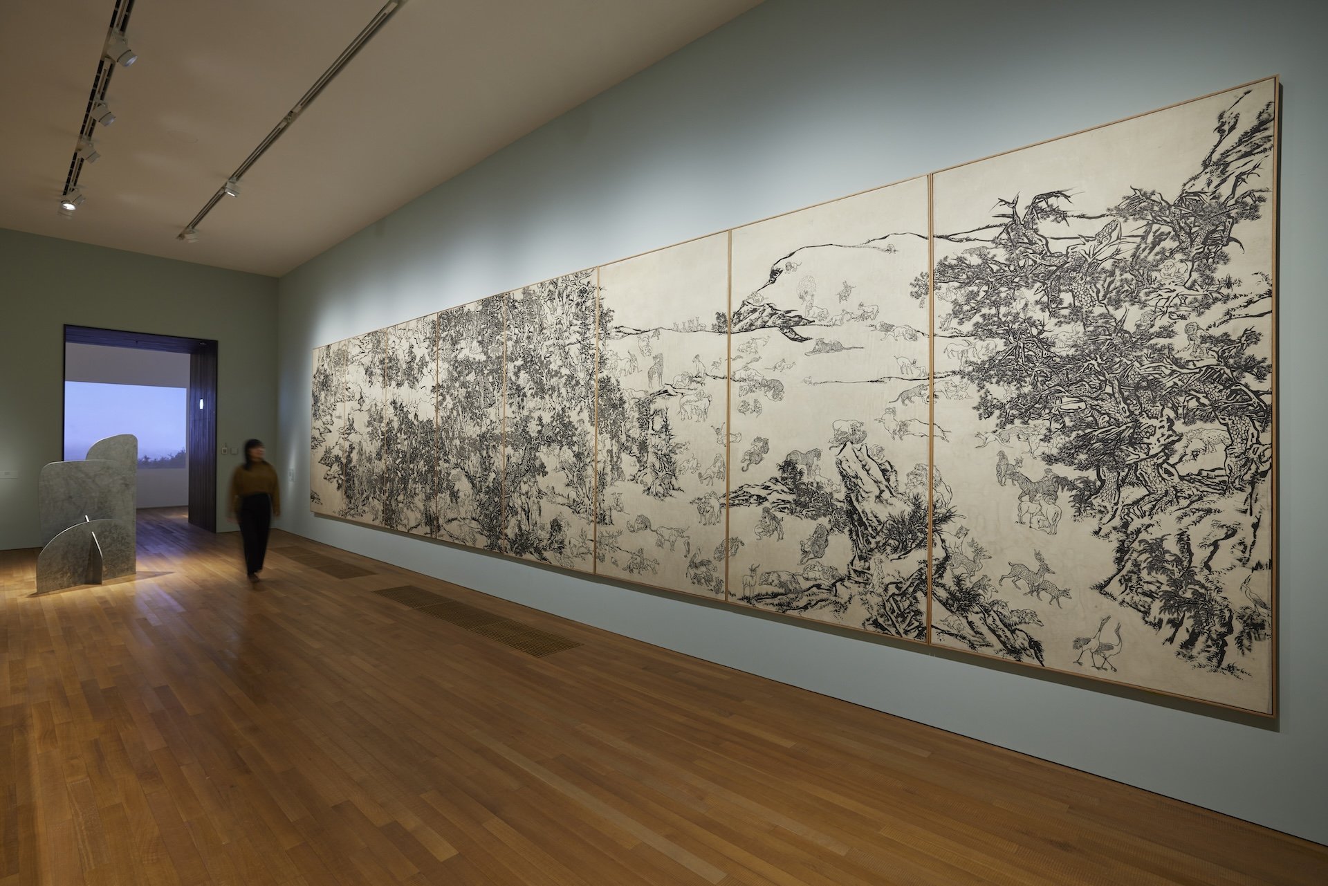A huge multi-panel black-and-white ink painting is mounted on canvas in a gallery room with grey walls and wooden flooring. The painting portrays humans and animals resting and interacting amid rocks and trees. They are mostly in pairs and have different poses.
